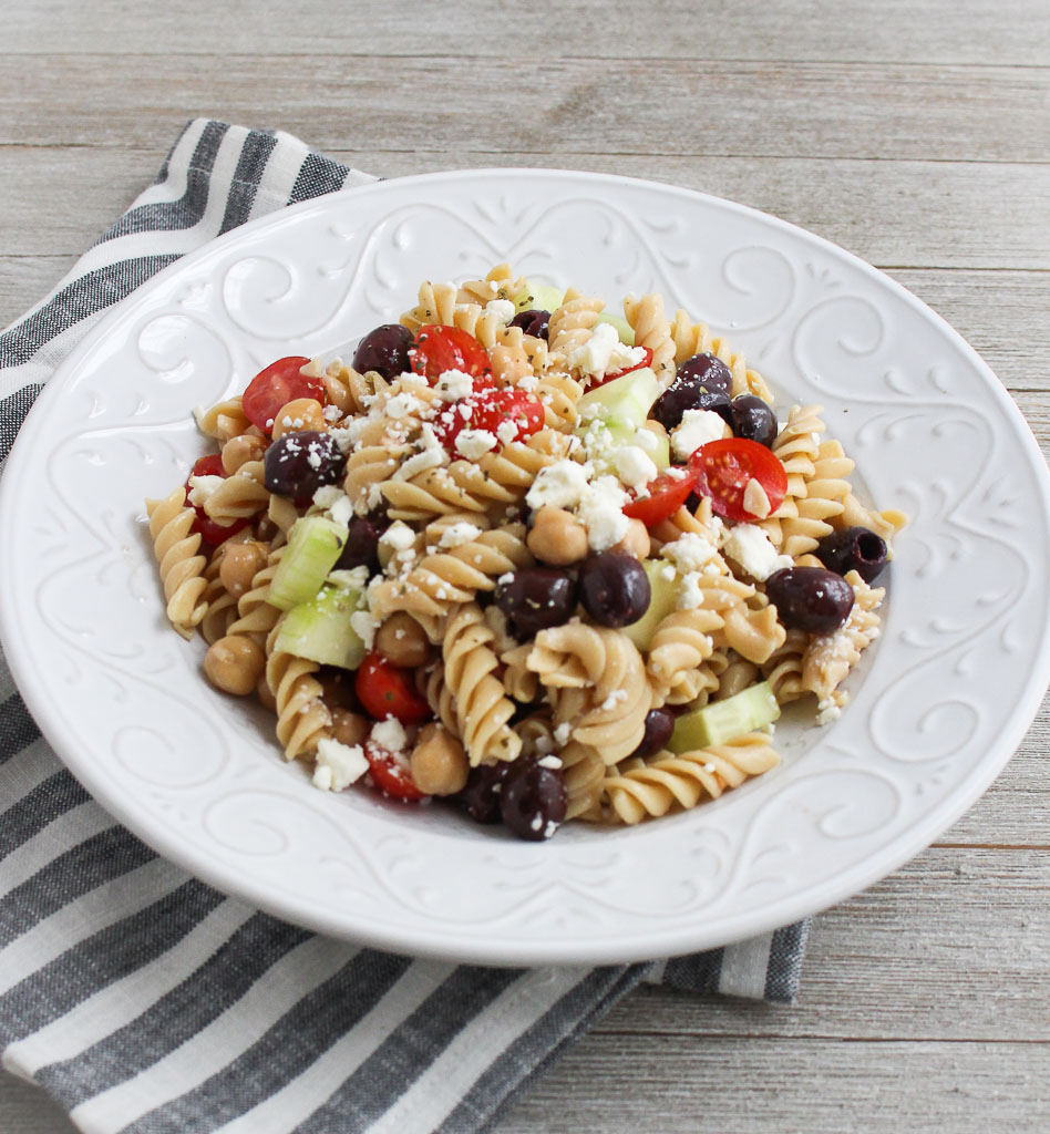 Greek pasta salad - Banza (chickpea) pasta salad with cherry tomatoes, cucumbers, olives, chickpeas, feta cheese, in a white dish